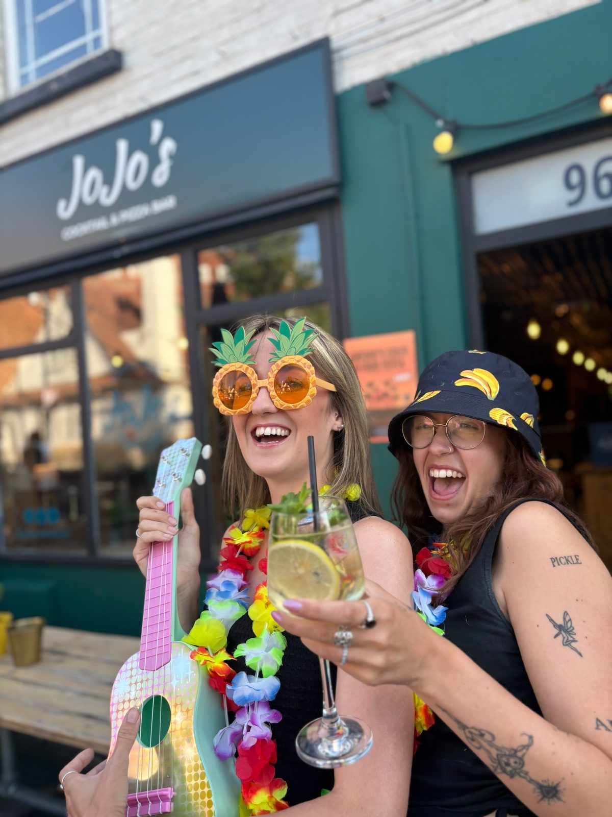Two people laughing outside a pub with colourful leis and a toy guitar
