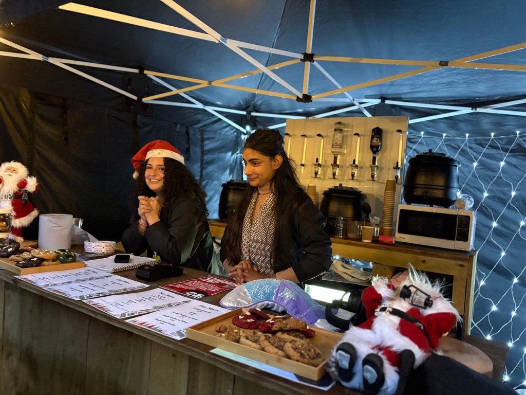 Two people behind a Christmas drinks stall with cookies, Santa decorations and fairy lights