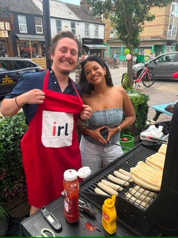 Two people smiling beside an outdoor barbecue, one wearing a red IRL apron
